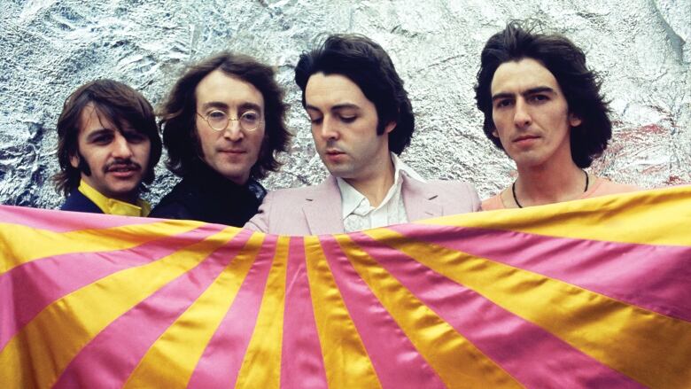 All four original Beatles members stand in a row, holding a flag that is stripes of pink and yellow.