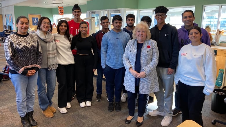 A group of teenagers stands smiling in a semi-circle around a middle-aged white woman with frosted blonde hair wearing a poppy.