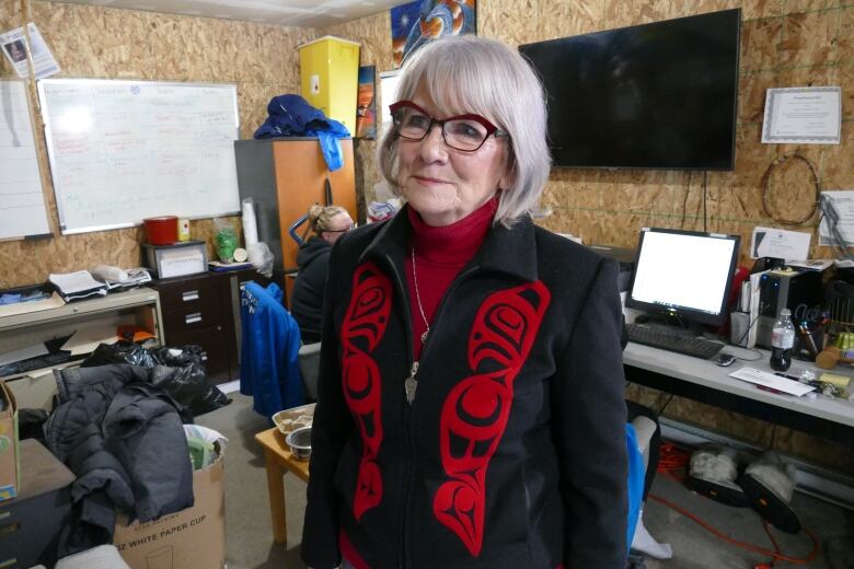 A woman is standing in a garage, with desks and computers behind her. There is a white board and other office supplies on the walls and tables.