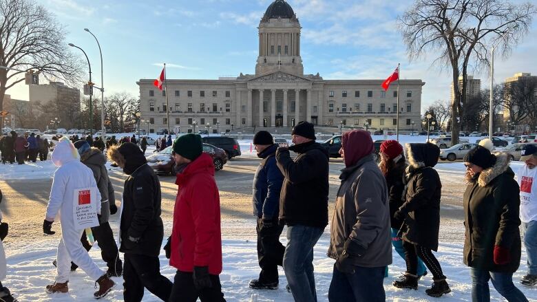 People walk across a snowy sidewalk, in front of the Manitoba Legislative Building that's across the street.