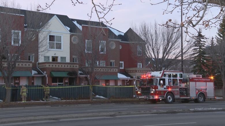 A wide shot of a townhouse surrounded by police tape with a  red and white fire truck parked in the corner.