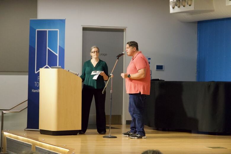 Two people stand on a stage. One is at a podium and one speaks into a microphone. 