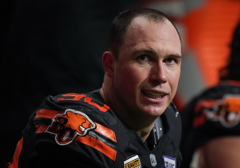 A football player in an orange and black uniform looks to his side, at the camera. His helmet is off.