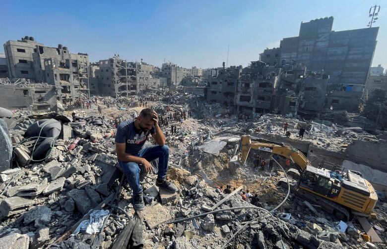 A man sits on a pile of rubble with his heads in his hands.