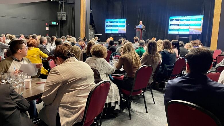 175 people sit at table during a conference as a man stands presenting at the front on a stage. 