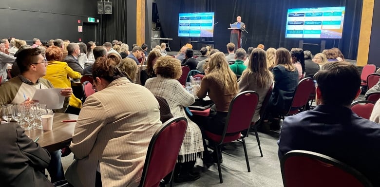 175 people sit at table during a conference as a man stands presenting at the front on a stage. 