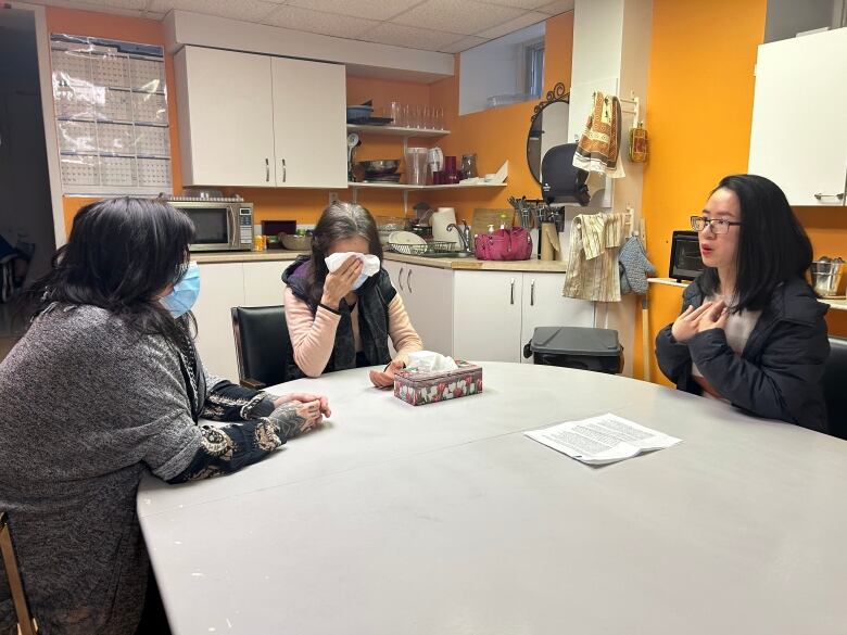 Three women sit around a table, one holding a tissue to her face. 