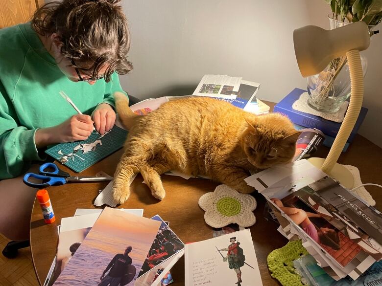 A person sitting at a table covered in paper they're cutting. There is also an orange cat on the table. 