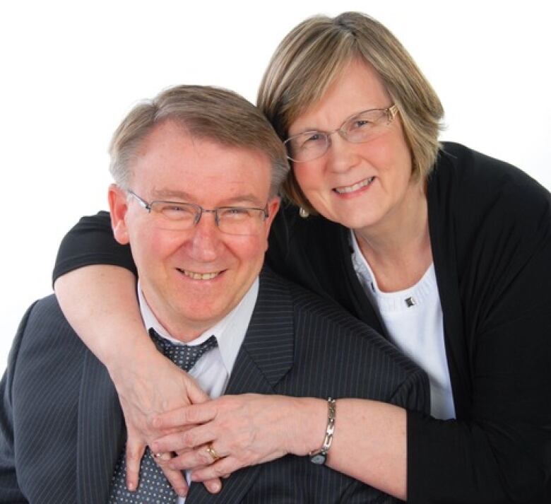 Man with grey hair and glasses sits as woman with grey-blonde hair and glasses hugs him around the neck from behind.