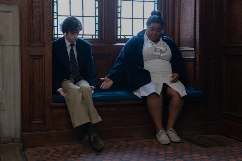 A young man in a school uniform sits next to a woman in a cafeteria gown. The woman holds out her hand to the young man, who is seated in way that suggests he is  nervous or uncomfortable. 