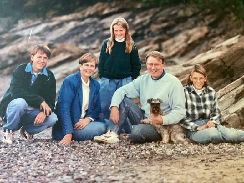 Man and woman sit with three children and a dog at a rocky beach.