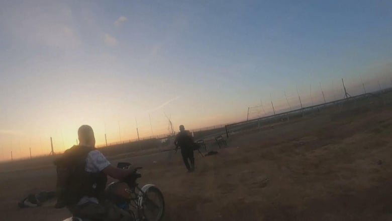 One man on a motorcycle and another on foot head toward a broken-down fence in dawn light.