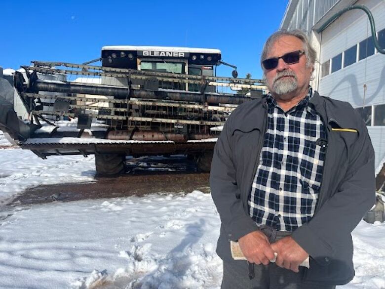 A man with a grey beard and hard wears a blue and white flannel shirt and a grey jacket. he stands in front of a combine harvester. There is snow on the ground. 