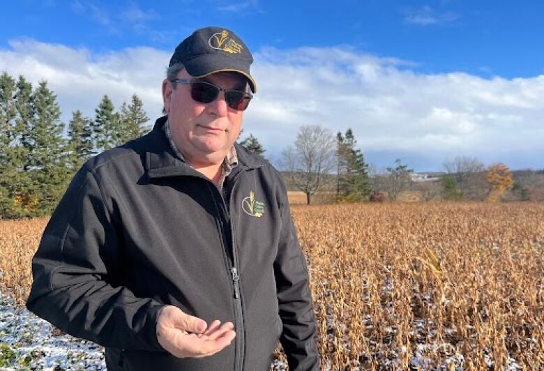 A man with black sunglasses wears a black hat and jacket. He stands in a feild with stocks of grain in the background. Patchy snow covers the gorund. 