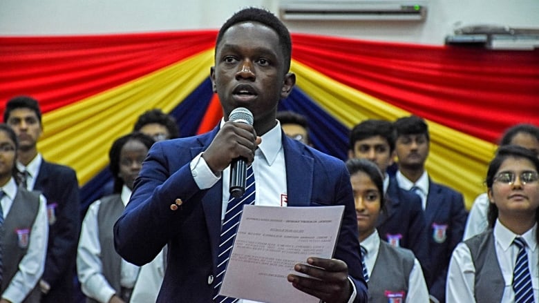 An older teen wearing a blue blazer and tie holds a sheet of paper while delivering a speech into a microphone. Numerous other teens standing behind him, all wearing school uniforms, listen attentively. 