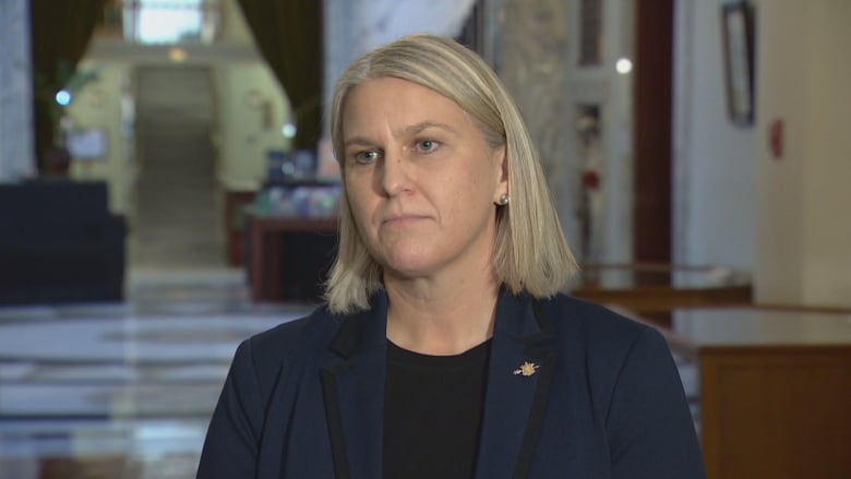 A woman with straight, bright blond hair in a bob looks off to the left of the frame. She's wearing a black shirt and a blue blazer.