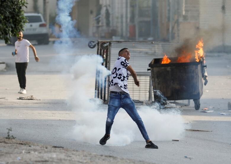 A man in a t-shirt and jean is shown in a throwing motion on an urban street. Nearby, a dumpster is on fire.