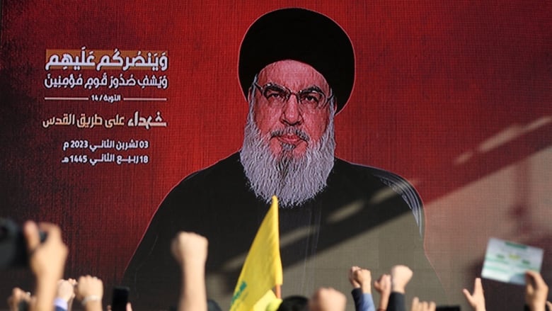 A bearded, bespectacled man in an head covering is shown projected on a screen in a public space with people raising fists in front of the screen.