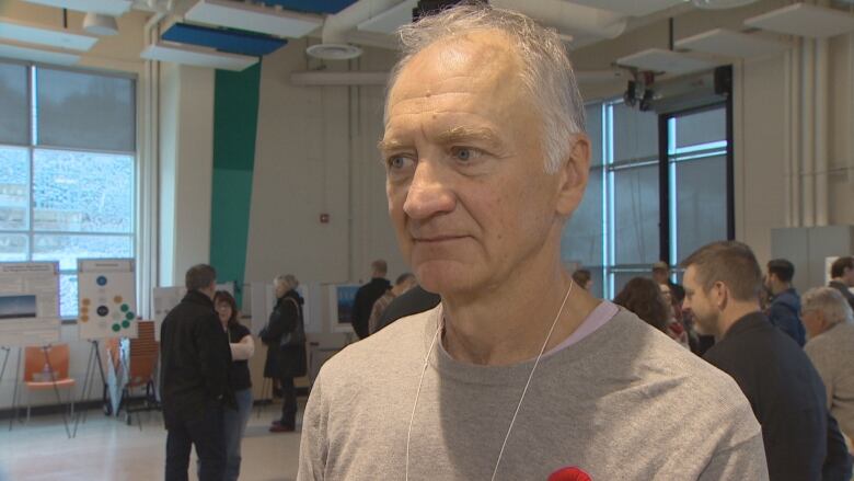 A man with white hair and grey t-shirt is standing in a room. The crowd mingles behind him.