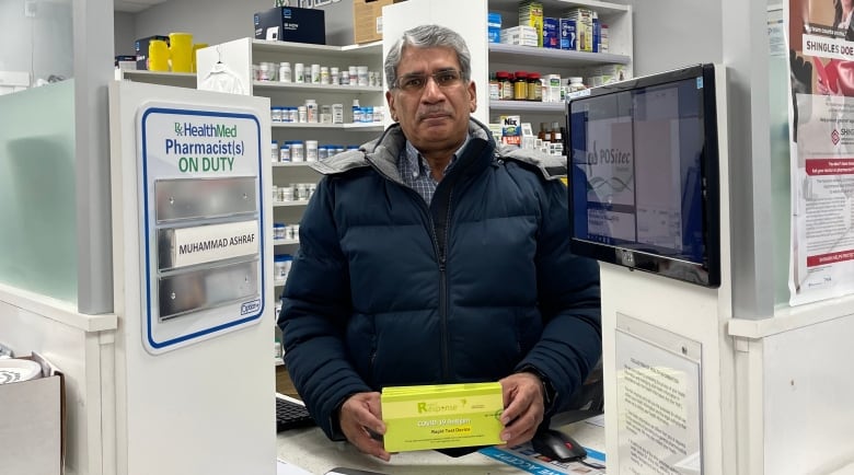 Pharmacist Muhammad Ashraf stands behind the counter at his pharmacy holding a light green box of rapid COVID-19 tests