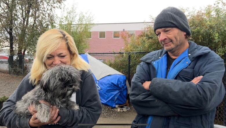 A middle-aged man and woman along with their small terrier mix dog standing at a homeless camp in early November.