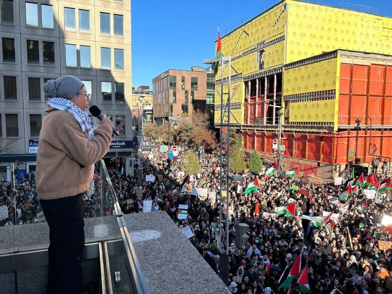 a woman in a grey hijab and a Palestinian keffiyeh around her neck addresses the crowd with a mic