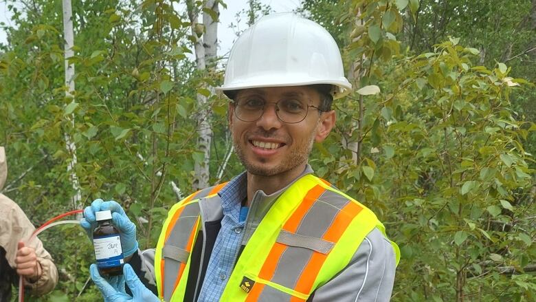 A man wearing a white helmet and neon yellow construction vest holds a bottle, smiling. He is standing outside with greenery behind him.