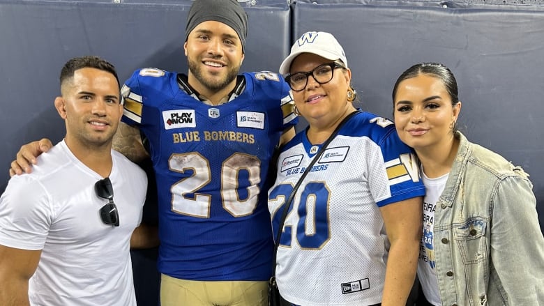 Four people stand should-to-shoulder beside one another. One, in the middle, wears a football uniform.