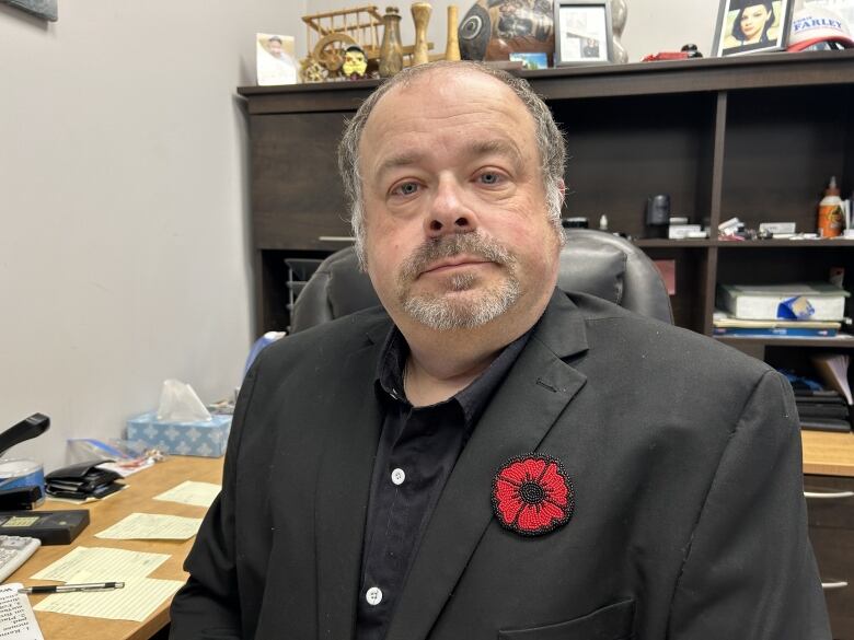 A man in a black dress shirt and a black jacket sits at a desk. The man is wearing a red poppy. 