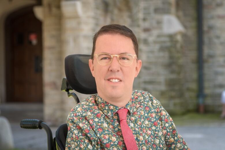 A man in a floral shirt and tie in a wheelchair looks into the camera. 