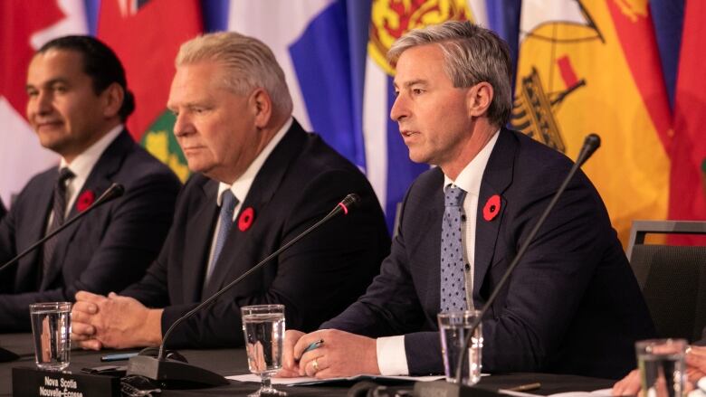 Three men wearing suits sit at a long table with microphones in front of them. Provincial flags can be seen behind them.