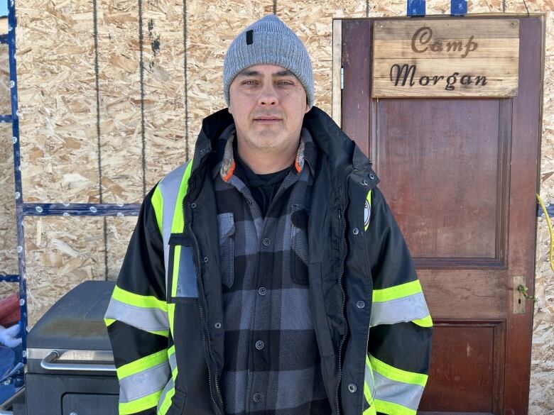 Man wearing a grey tuque stands wearing an open jacket with reflective stripes. A sign behind him says Camp Morgan  