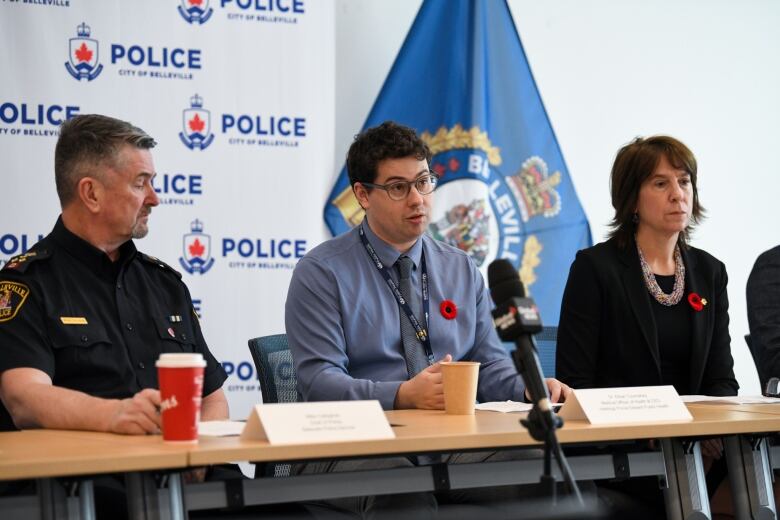 Three people are sitting at a table in front of microphones. One is in a police uniform, the centre one has glasses and a tie and the woman sits to his left. In the background is the city of Belleville's flag.