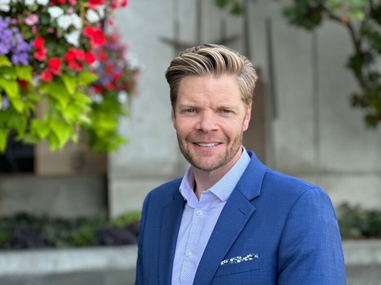 A white man with blond hair smiles into the camera. He is wearing a blue button-up shirt and a blue blazer with a pocket square. 