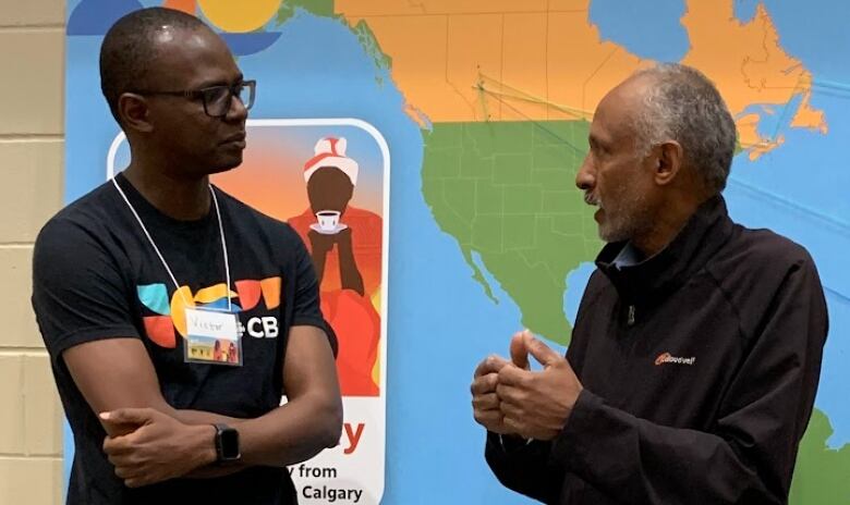 A young man listens as a man with grey hair talks in front of a large map.