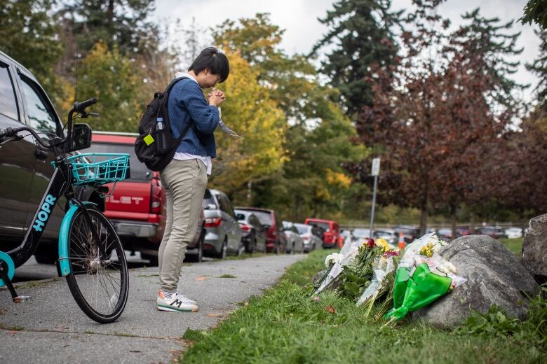 A person folds their hands next to a lawn with flower bouquets that have been placed on a rock.