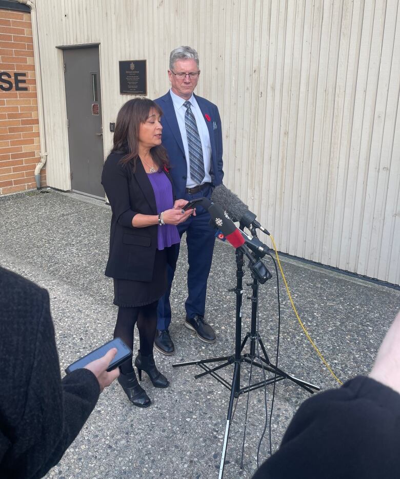 A woman reads from her phone to a series of standing mics, next to a man in a suit.