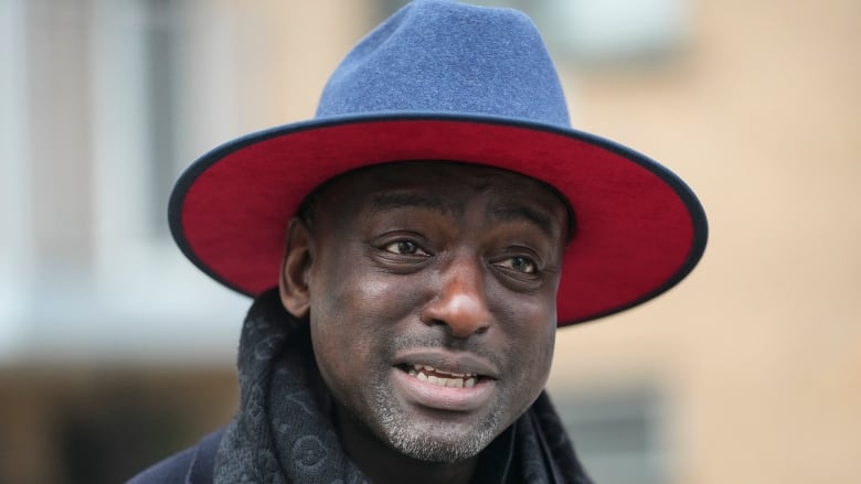 A dark-skinned man in a hat is seen in a closeup in an outdoor photo.