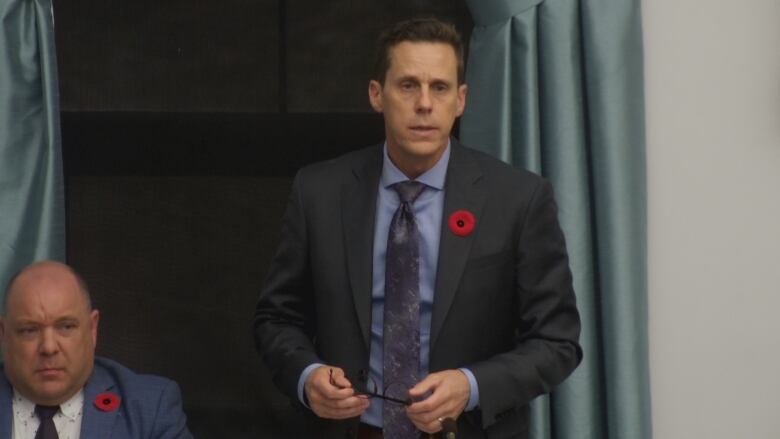 A man in a dark suit and blue tie stands while taking questions from politicians.