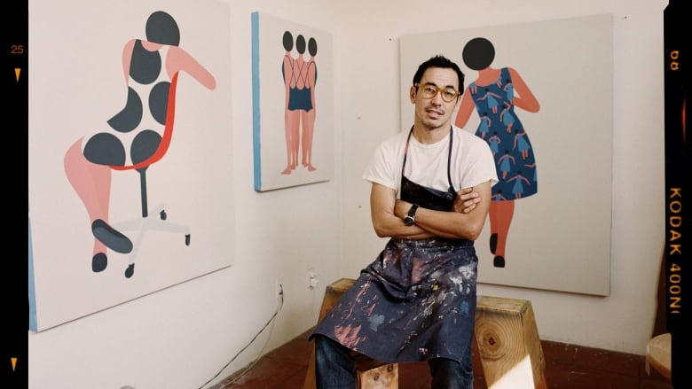 Portrait of the artist Geoff McFetridge. He is a man with short hair and a goatee. He wears dark frame glasses, a white T-shirt, jeans and a paint-splattered apron. He is seated on a wooden bench with his arms crossed. Three of his paintings hang on the white walls behind him. They are minimalistic representations of female figures with their backs to the viewer.