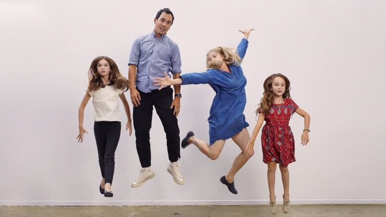 Photo of a family, jumping in the air against a white wall: two young girls flank their parents at centre: a dark-haired man and a blonde woman.