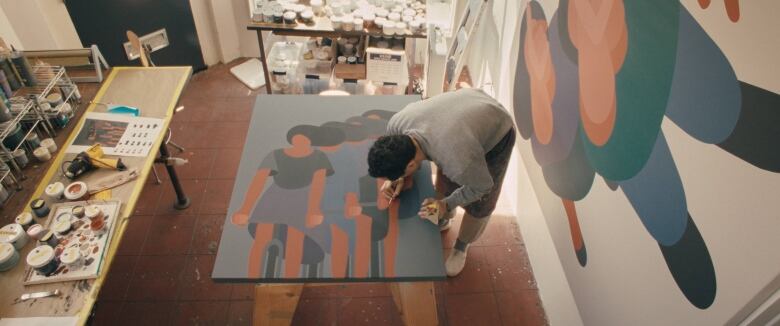 Photo of an artist, Geoff McFetridge, painting in a sun-filled studio. He is photographed top down. He is bent over a canvas, painting. Another large painting of figures in the same minimalist style hangs on the wall behind him. Surrounding his work surface are two more long tables covered with art supplies.