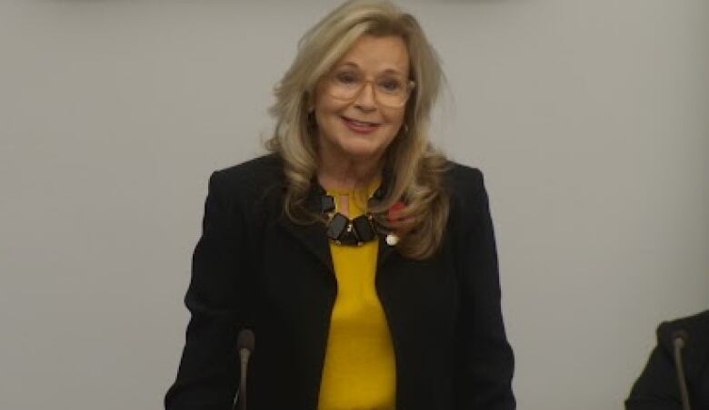 Woman with glasses and long blonde hair in dark blazer and yellow blouse. 