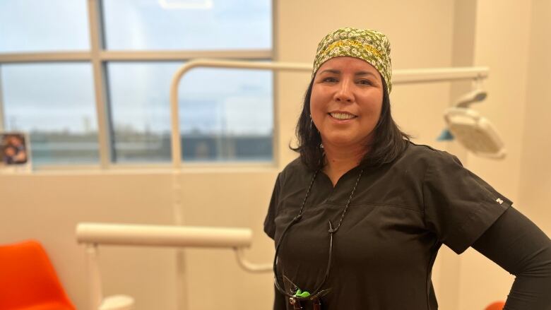 A woman can be seen wearing black scrubs, she has glasses hanging around her neck. She is smiling at the camera. 