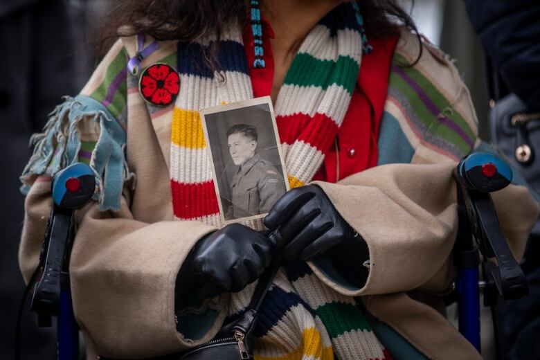 A woman sitting in a wheelchair holds a black and white photo of a soldier. She is wearing black gloves, a beige jacket, colourful scarf and a beaded poppy. 