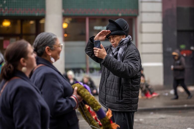 A man in a black jacket, toque and baseball hat is holding a phone in one hand and saluting with the other. In the foreground are two women holding a wreath with a purple ribbon. 