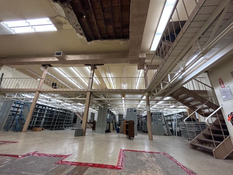 A large room is filled metal shelves, with stairs leading to a mezzanine level. There's a large hole in the ceiling.