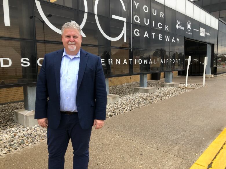 A man with white hair and a white goatee wearing a blue suit and jeans standing in front of a glass sign