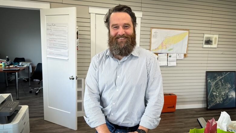 A man with brown hair and a bushy beard and white shirt stands behind a counter in an office.