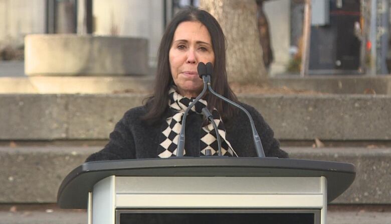 a woman with black hair and a black coat speaks into a microphone